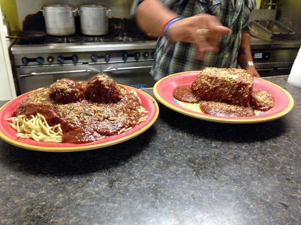 Two plates of spaghetti and rectangular meatloaf with sauce, in a kitchen setting; person gestures.