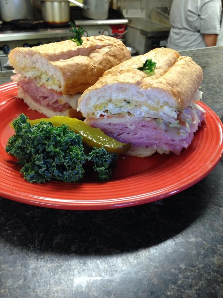 A sliced sandwich with ham, cheese, and veggies on a red plate, with a pickle and kale.