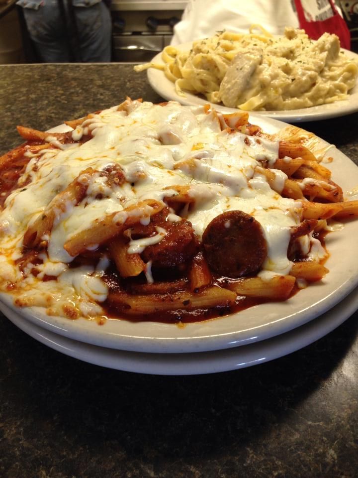 Pasta dish with melted cheese, meatballs, and sausage on a white plate. Another pasta dish in the background.