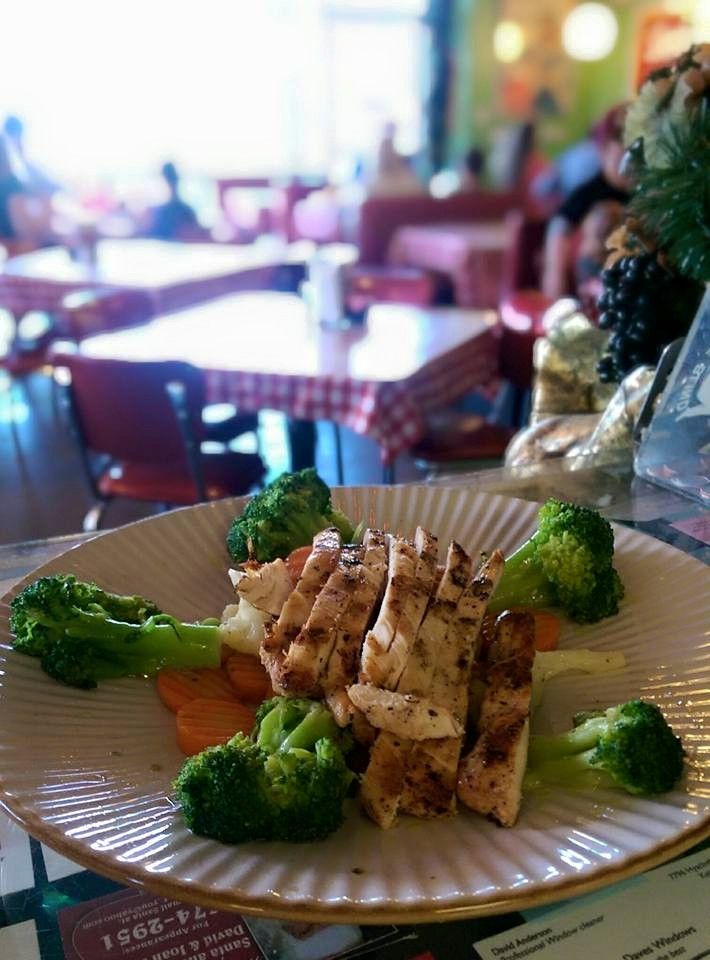 Grilled chicken and broccoli on a plate at a restaurant, with a blurred background of tables and people.