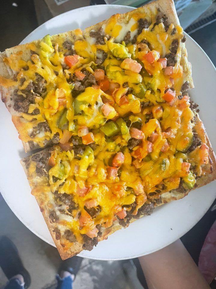 Square slice of bread topped with ground meat, cheese, diced tomatoes, and avocado on a white plate.