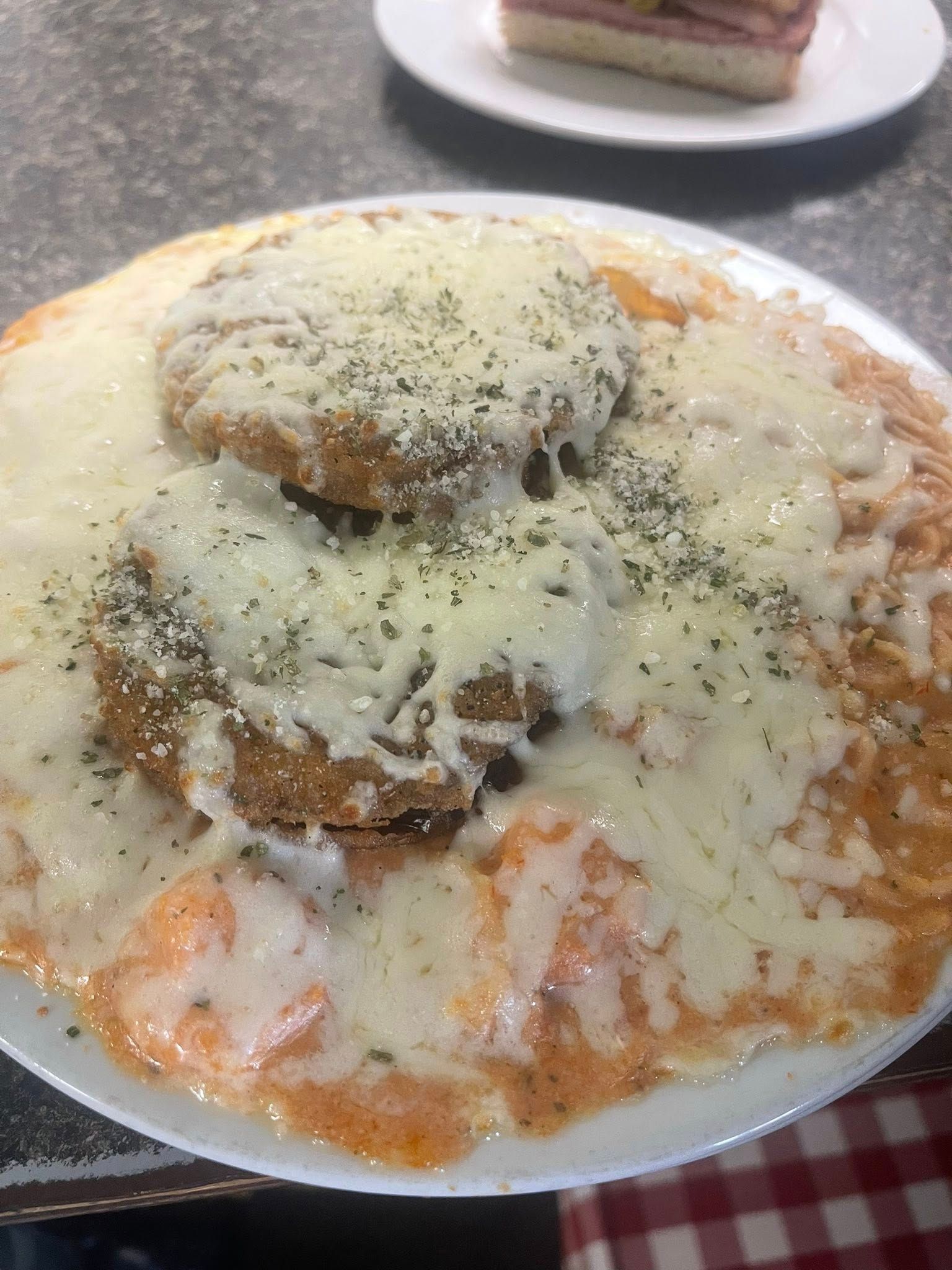 Eggplant parmesan on a white plate, covered in sauce and cheese, with a sandwich in the background.