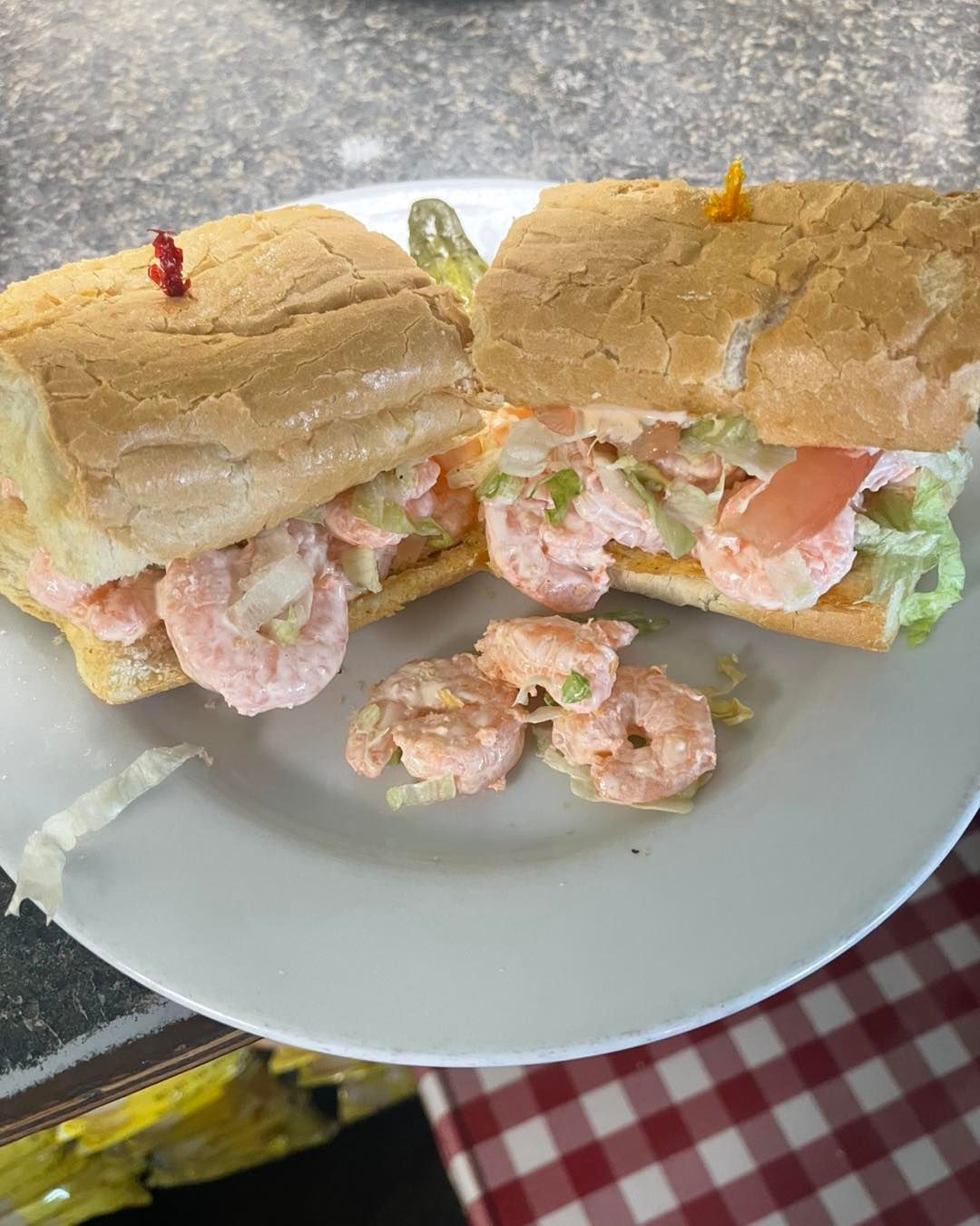 Two halves of a shrimp po' boy sandwich on a white plate, with some loose shrimp, and checkered tablecloth in the background.