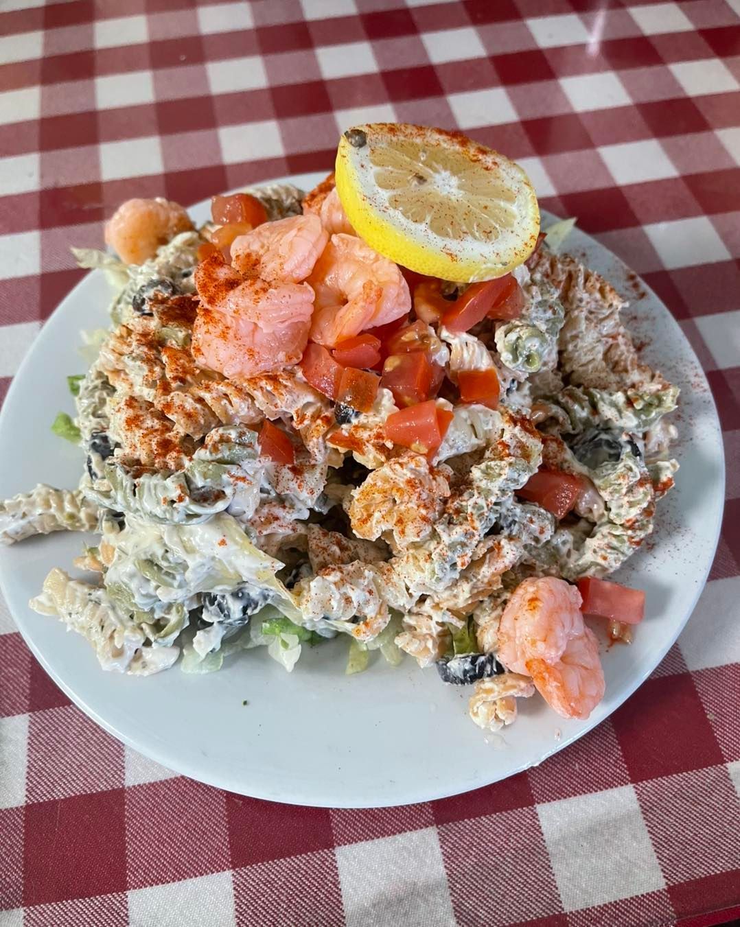 Pasta salad with shrimp, tomatoes, lemon on a white plate, red-checkered tablecloth.