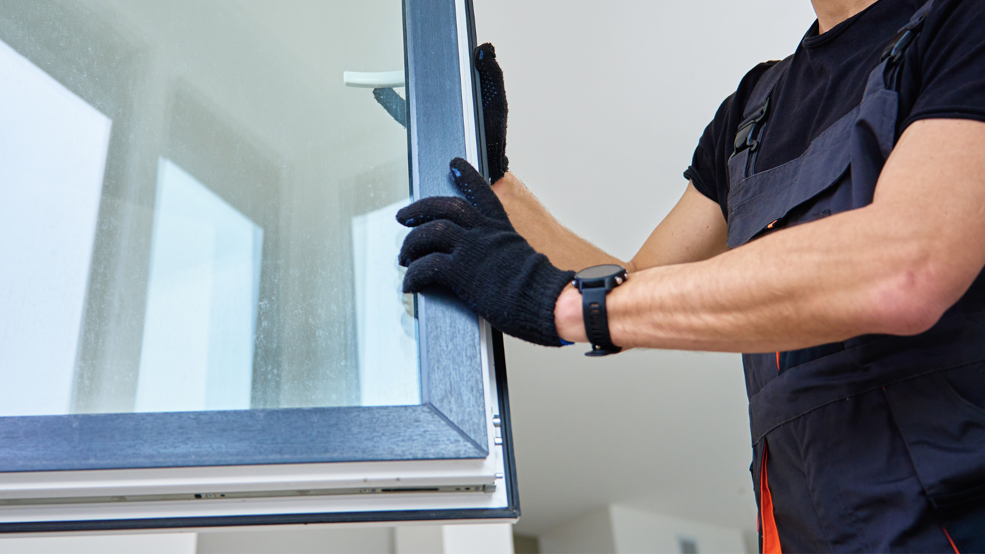 A person in work coveralls and black gloves installs or holds a modern grey-framed window.