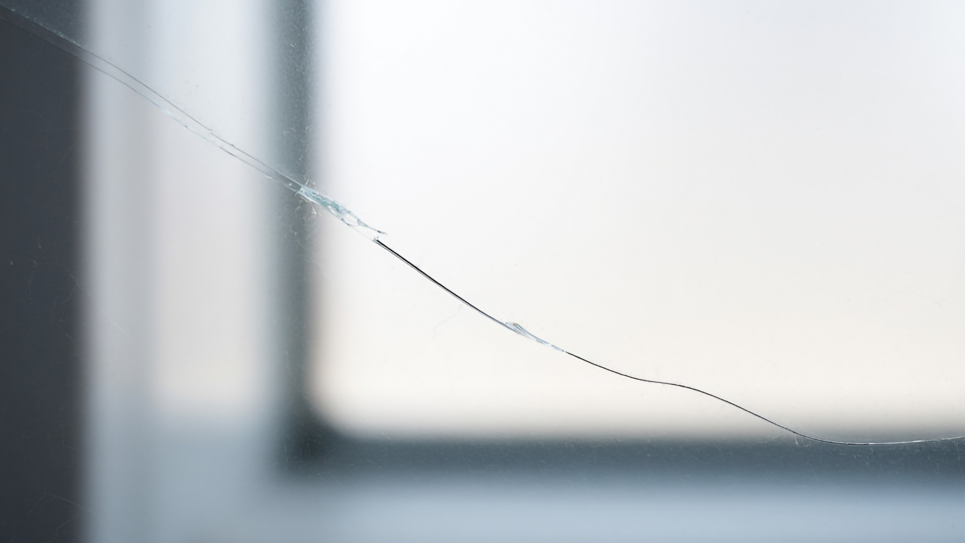 A close-up of a thin, jagged crack running diagonally across a glass surface, set against a blurry, light-colored background.