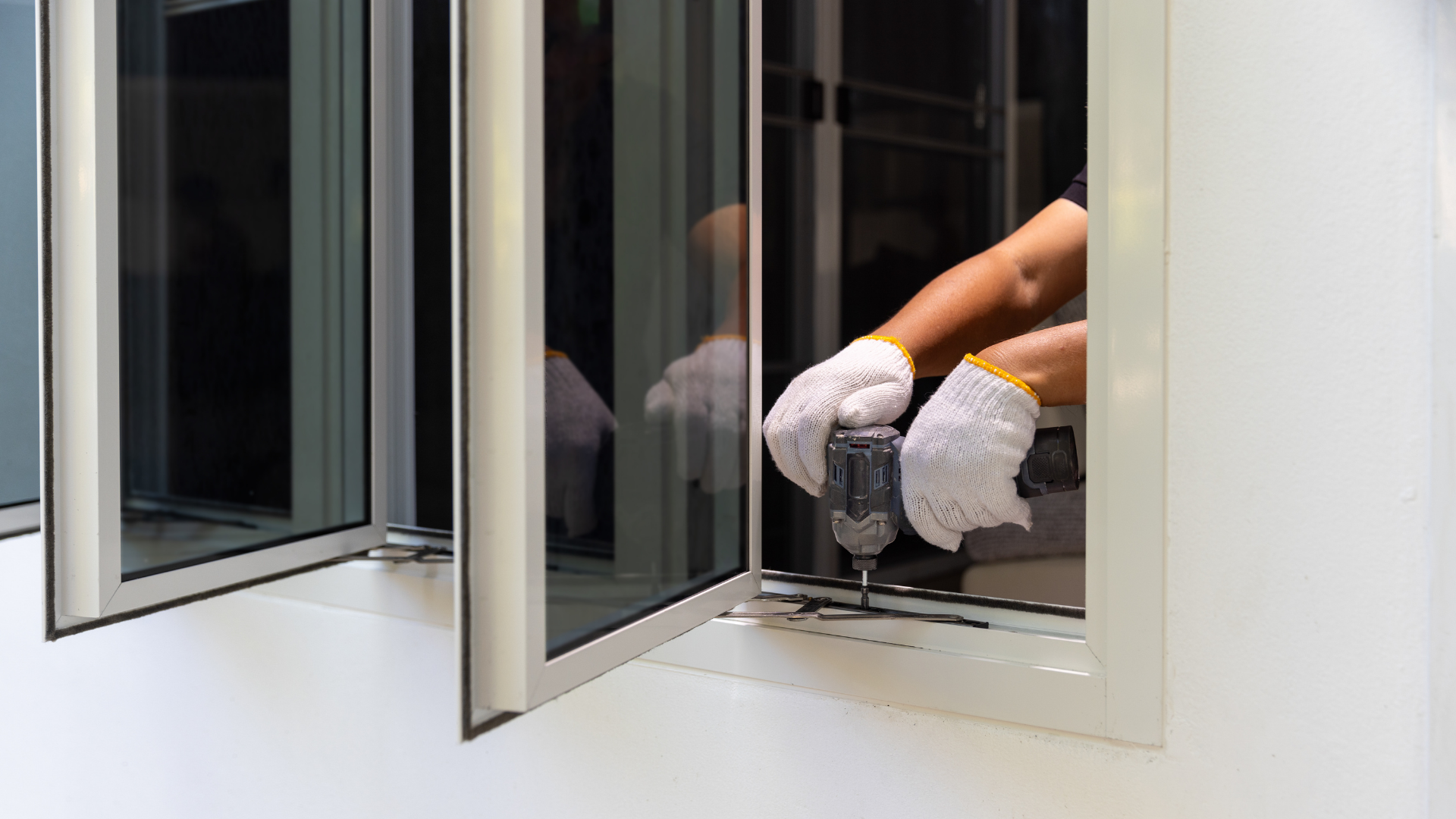 A person in white work gloves uses an electric drill to install or repair a white aluminum window frame.