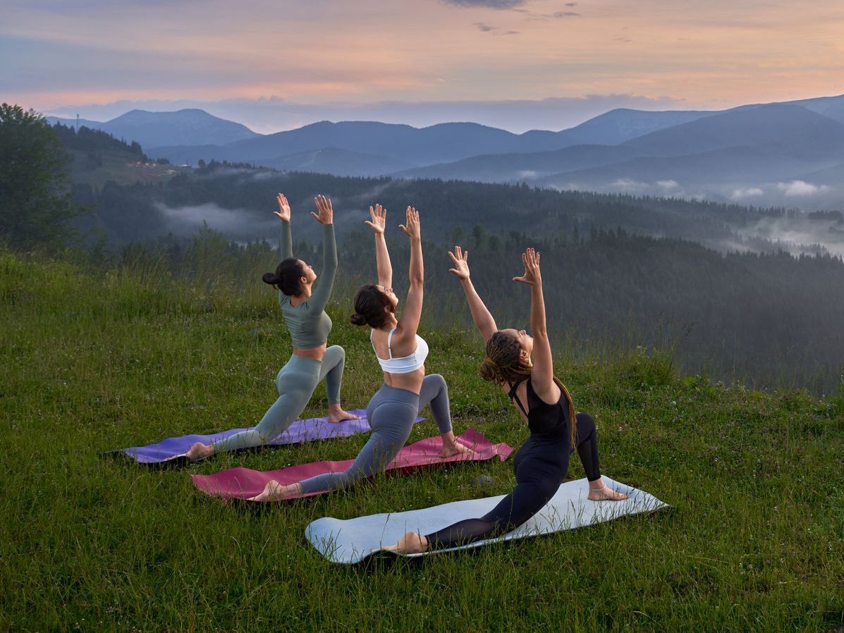 Three women are practicing yoga on top of a grassy hill.
