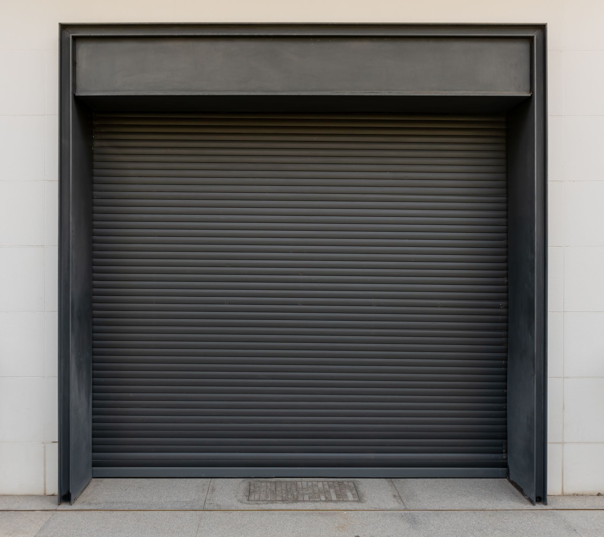 Closed, dark gray rolling security shutter framed in dark metal, set in a light gray wall.