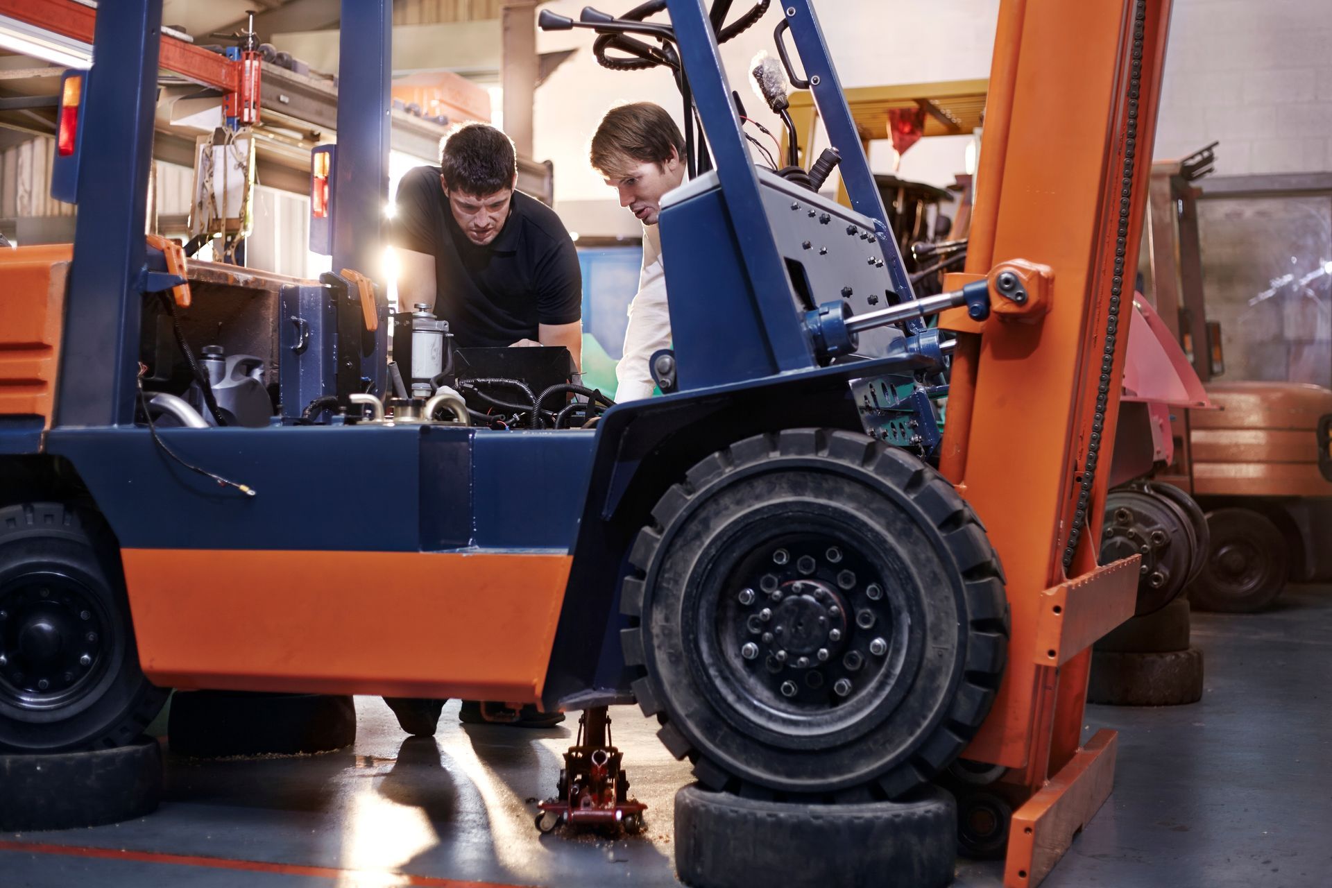 Two people working on a forklift in a garage; blue and orange colors. One is using a light.