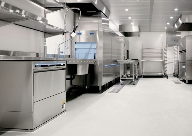 Bright stainless steel commercial kitchen with counters, ovens, and shelves in a clean white room