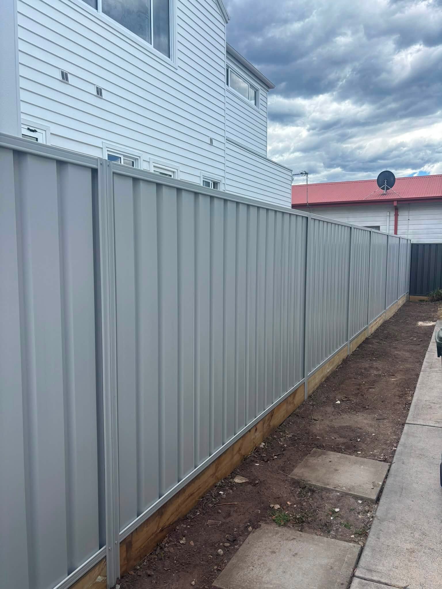 Gray metal fence beside a narrow dirt strip and sidewalk under a cloudy sky.