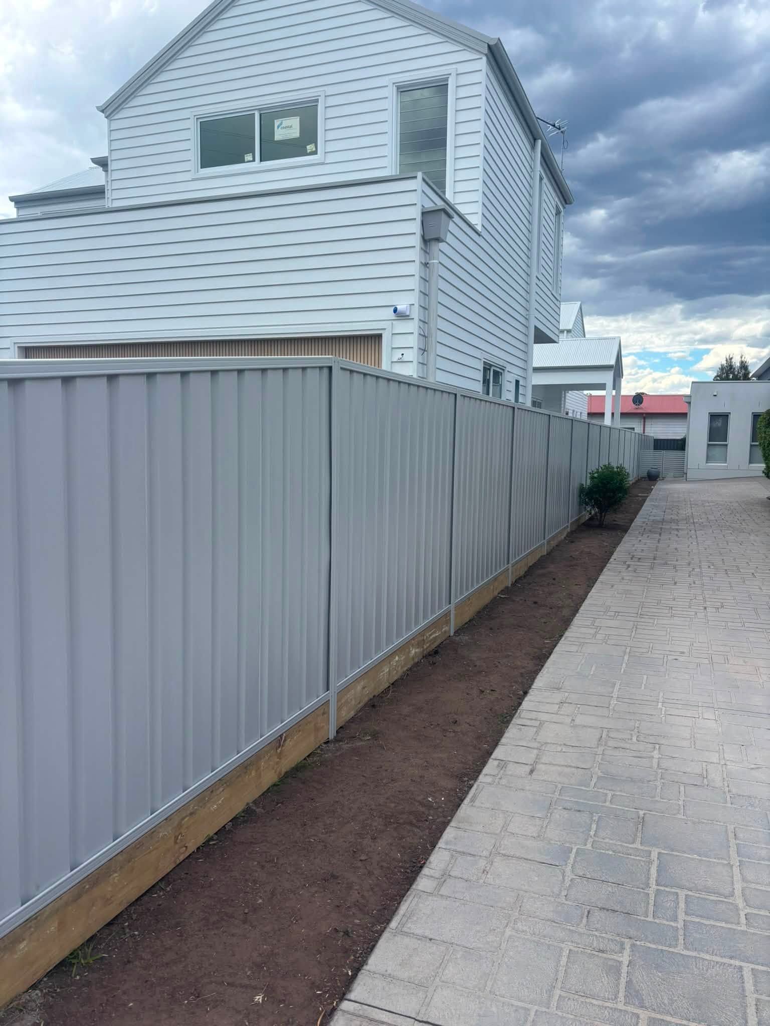 Narrow outdoor walkway beside a gray house and fence, with a paved path and dirt strip along the side.