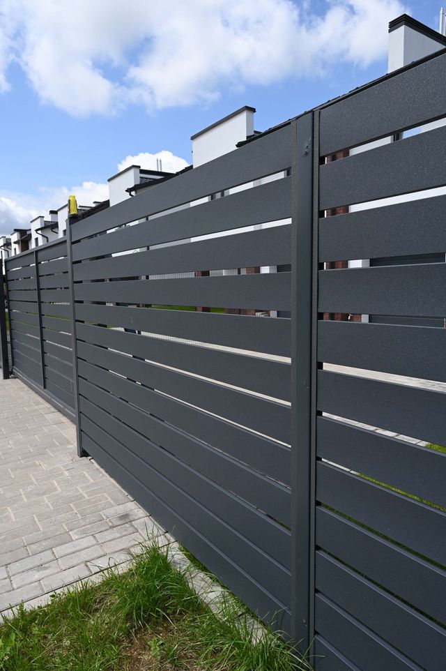 Dark gray slatted fence along a paved sidewalk under a partly cloudy sky
