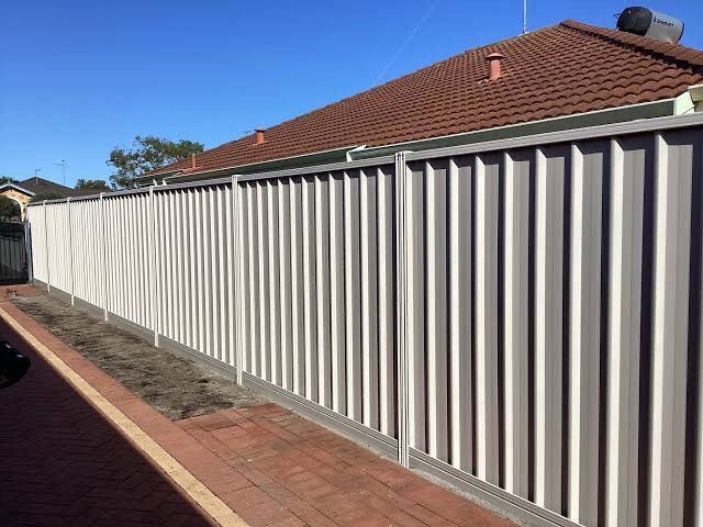 Long white metal fence beside a brick sidewalk and red-roofed house on a sunny day