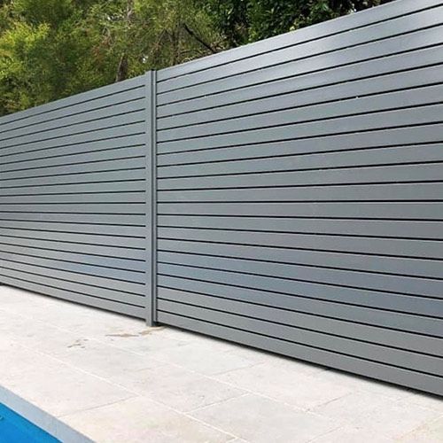 Gray slatted privacy fence along a tiled patio beside green trees and a blue pool edge