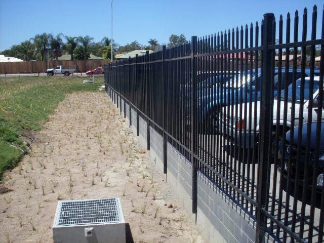 Rocky path beside a black metal fence with parked cars on the right and a grassy area on the left
