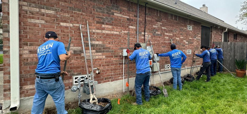 A team of men in blue shirts works along a brick wall, likely installing utilities, in a residential setting.