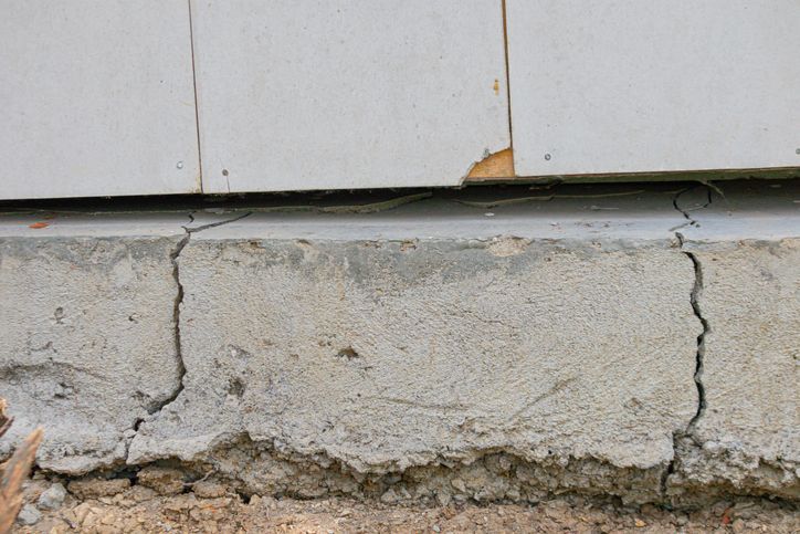 Close-up of a concrete foundation with vertical cracks. The foundation supports light-colored siding with a small piece missing.