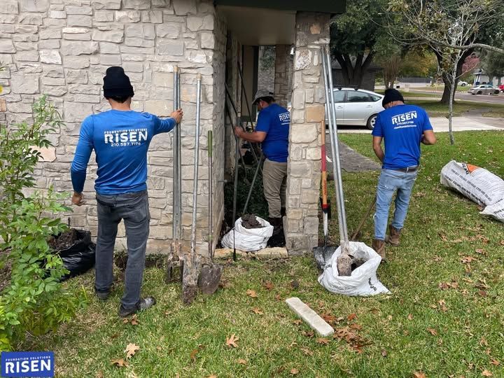 Three construction workers in blue shirts are working on a stone building exterior. They are using tools and stand in front of an open doorway.