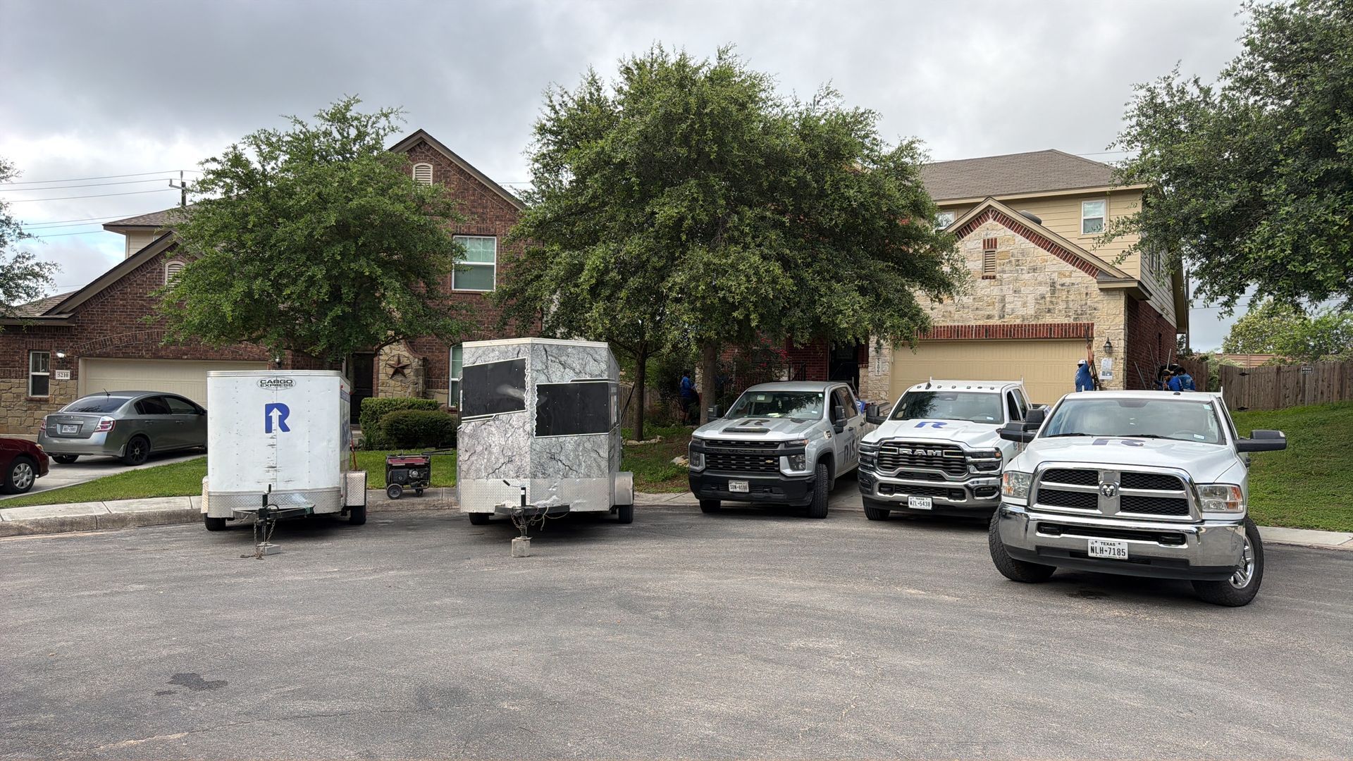 White trucks and utility vehicles parked in a residential driveway with two trees in front of houses.