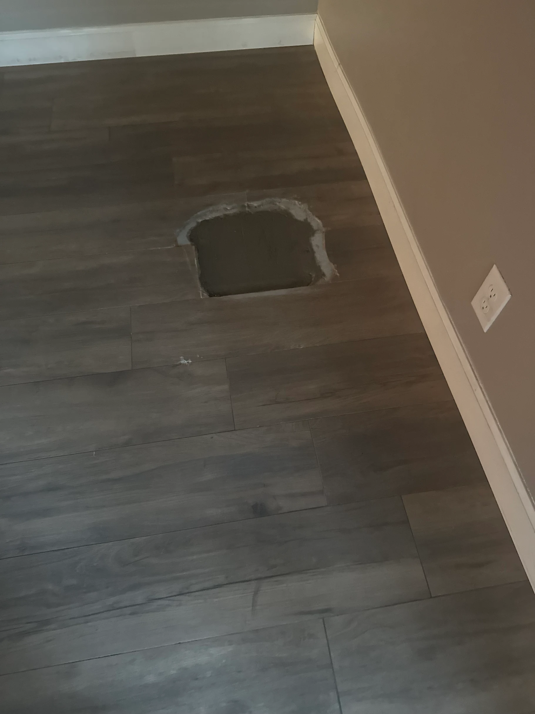 Hole in grey flooring, likely for access to plumbing. Light-colored walls and baseboard surround. Hole in grey flooring, likely for access to plumbing. Light-colored walls and baseboard surround.
