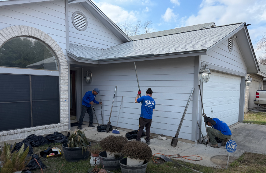 Three workers in blue shirts use long-handled tools to clean the exterior siding of a suburban house.