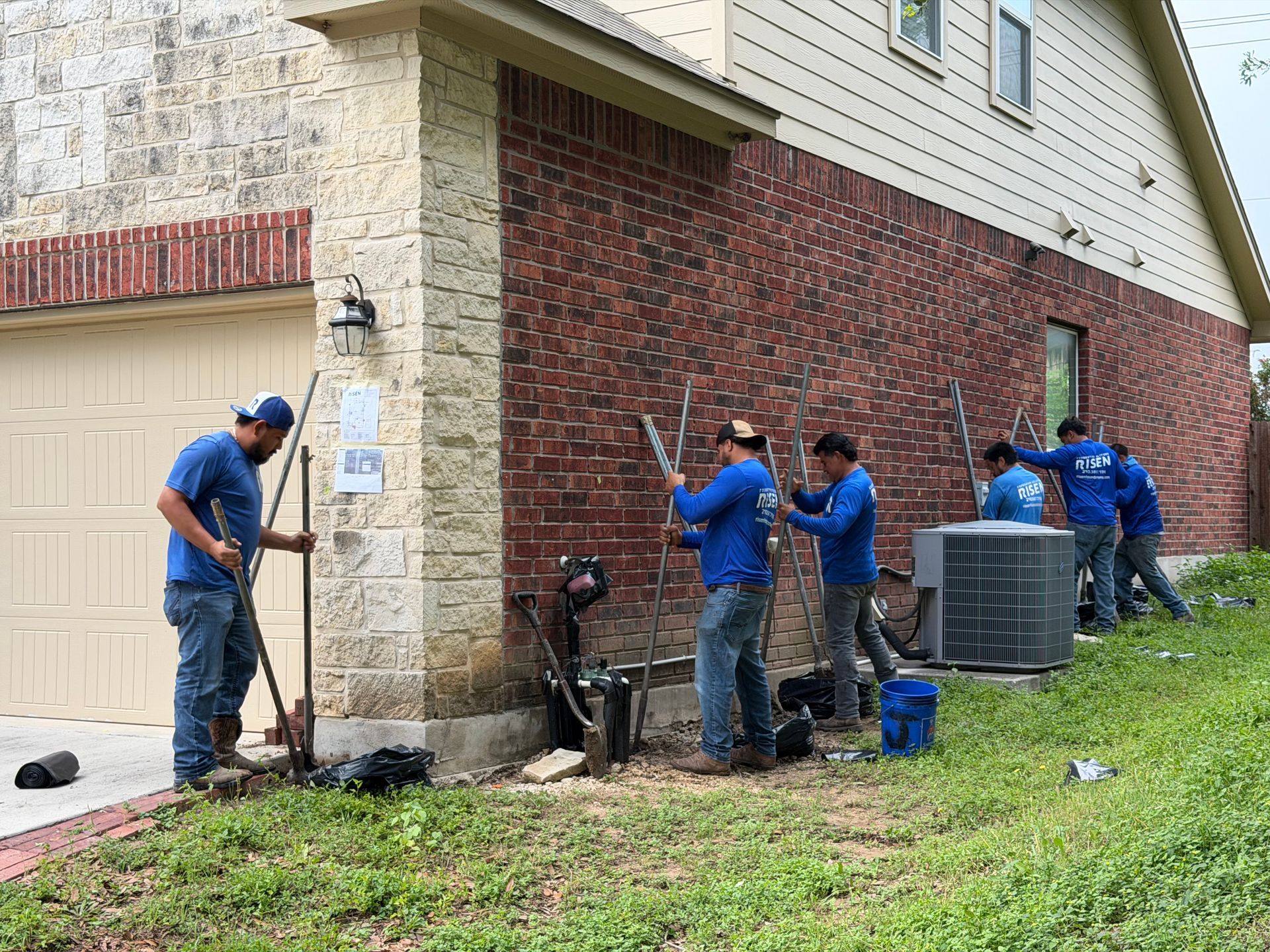 Workers repair an outdoor brick wall beside a house, with tools and an air conditioner unit nearby