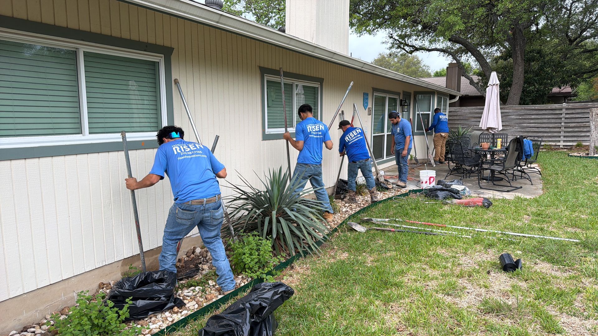 Workers landscaping along the side of a beige house, planting and trimming garden beds. Workers landscaping along the side of a beige house, planting and trimming garden beds.