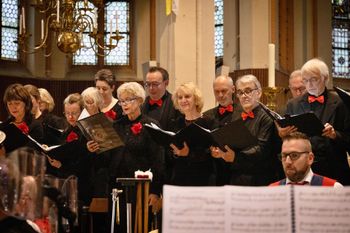 Een koor zingt in een kerk; de leden dragen zwarte kleding met rode strikjes en bloemen. Muziekstandaards.