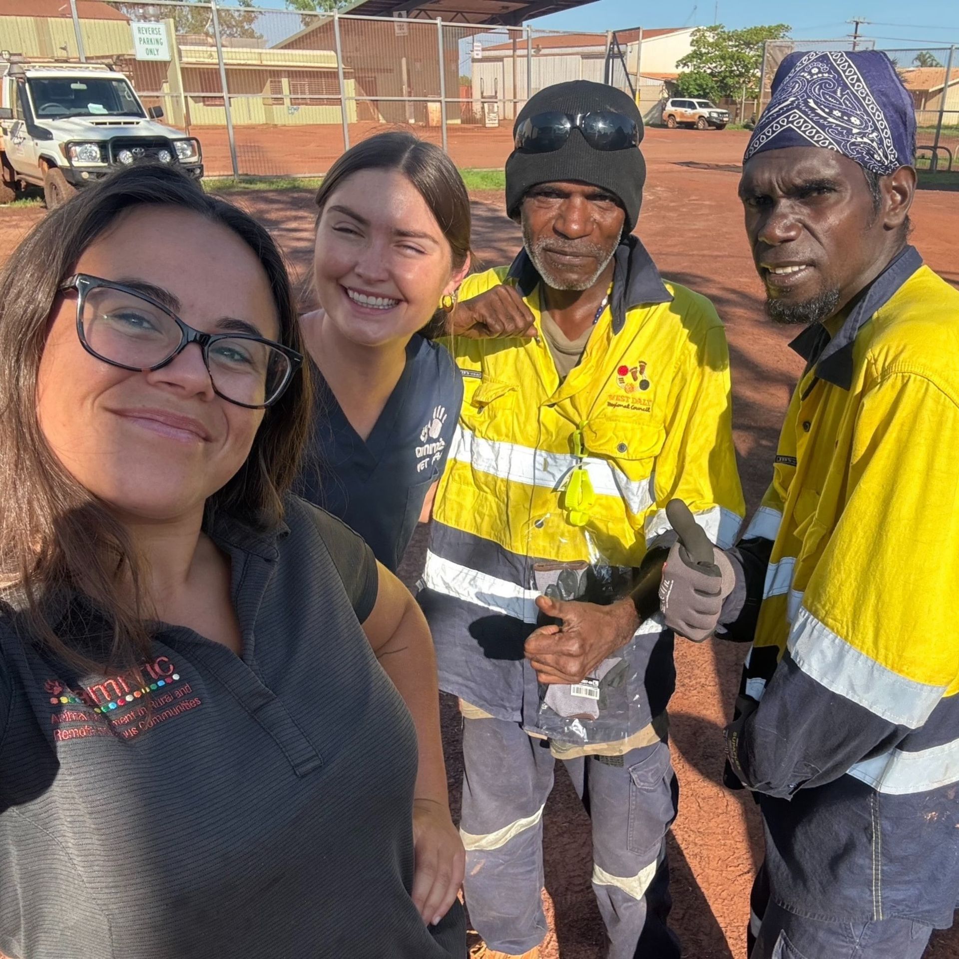 Four people smiling for a selfie outdoors. Two women, two men in work clothes. Sunny, red dirt ground.