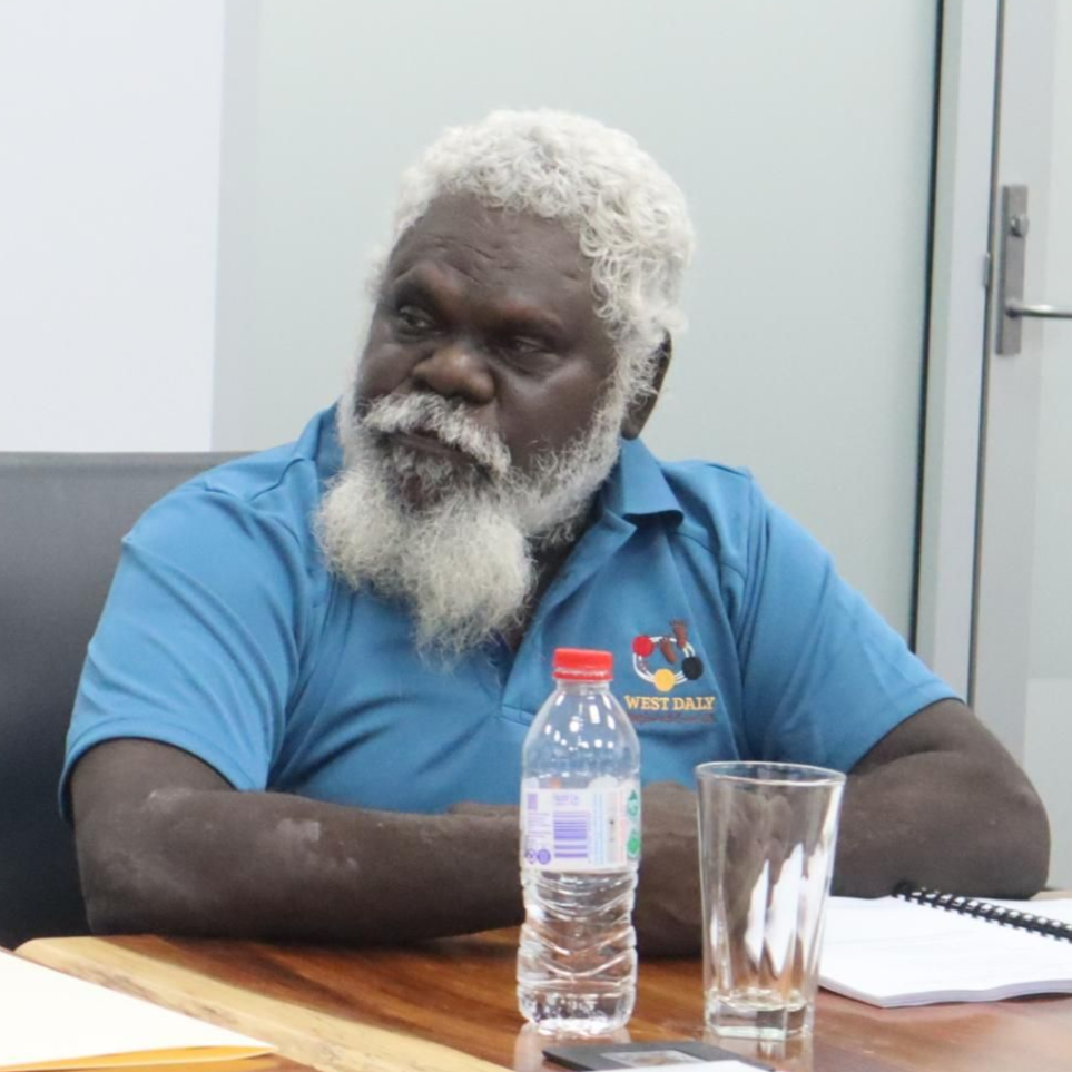 Man with white beard, blue shirt, seated at a table, looking thoughtful. Bottle of water and glass present.