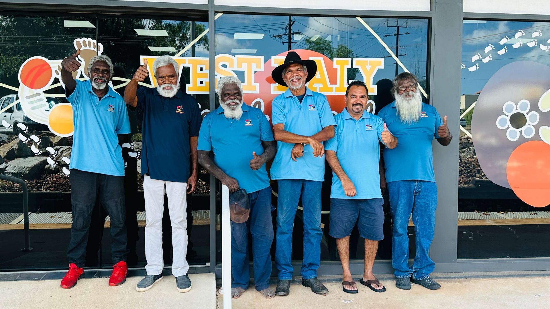 Group of men in blue shirts pose in front of a storefront with signage. Some raise fists or give thumbs up.