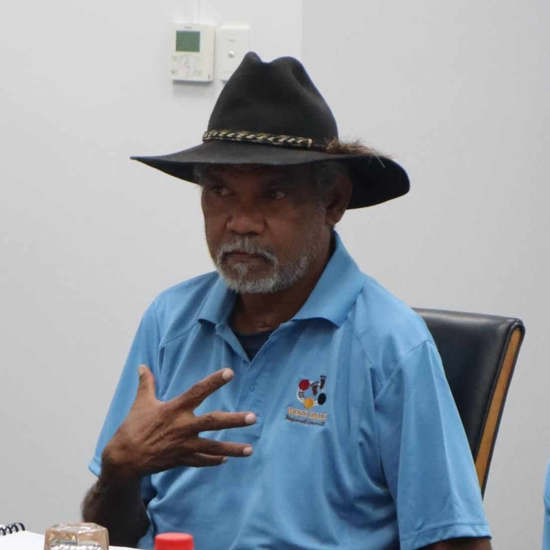 Man wearing a black hat, blue shirt, gesturing with his hand. Sitting indoors, light-colored walls.