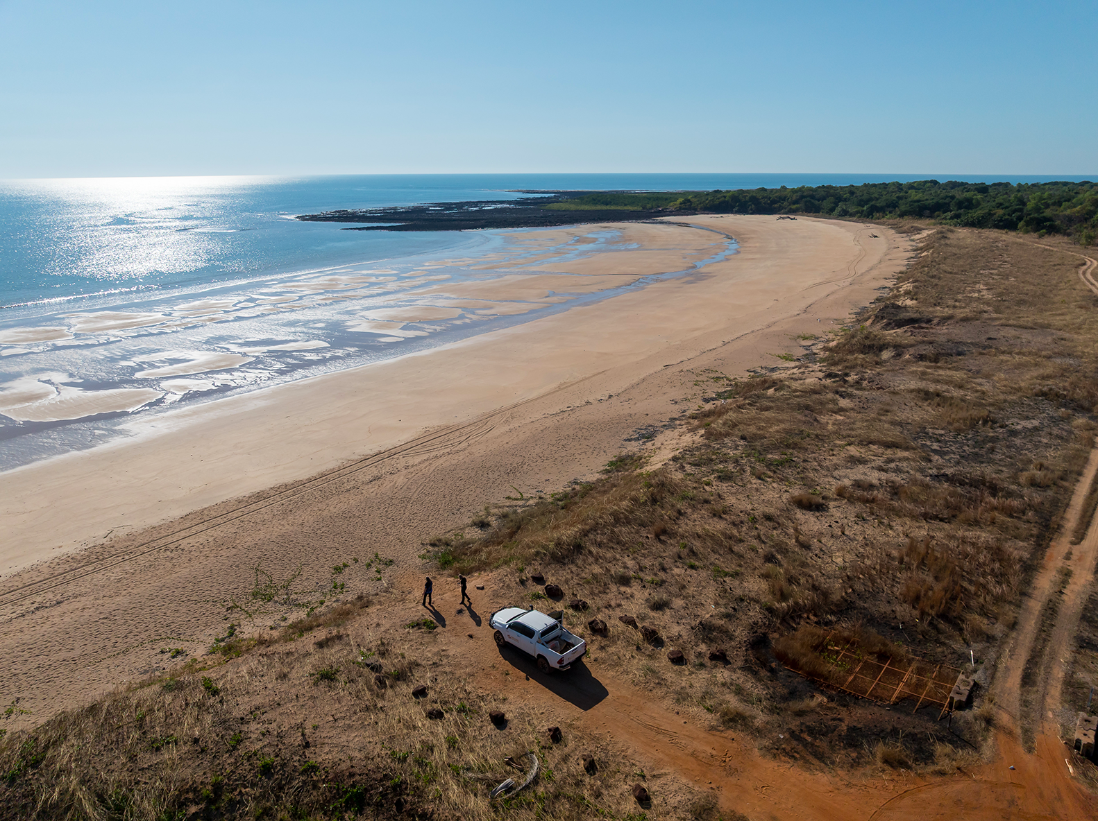 White truck on a sandy beach. Ocean and coastline in the background, blue sky.