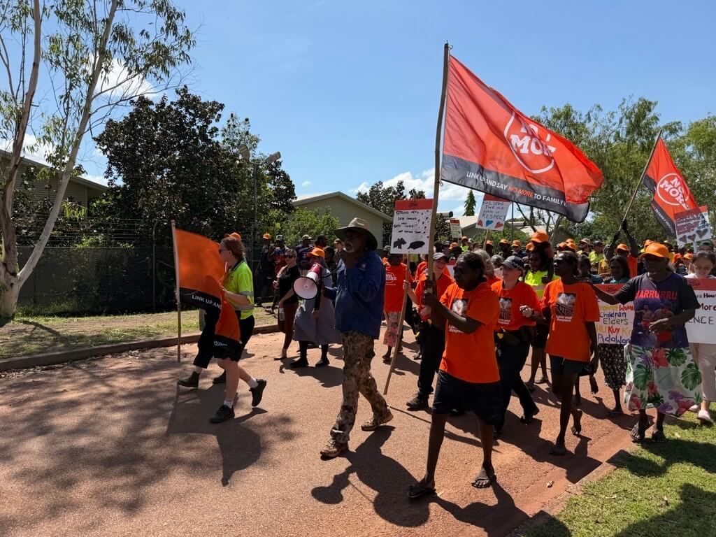 People marching and holding signs and flags. Many are wearing orange, under a blue sky.