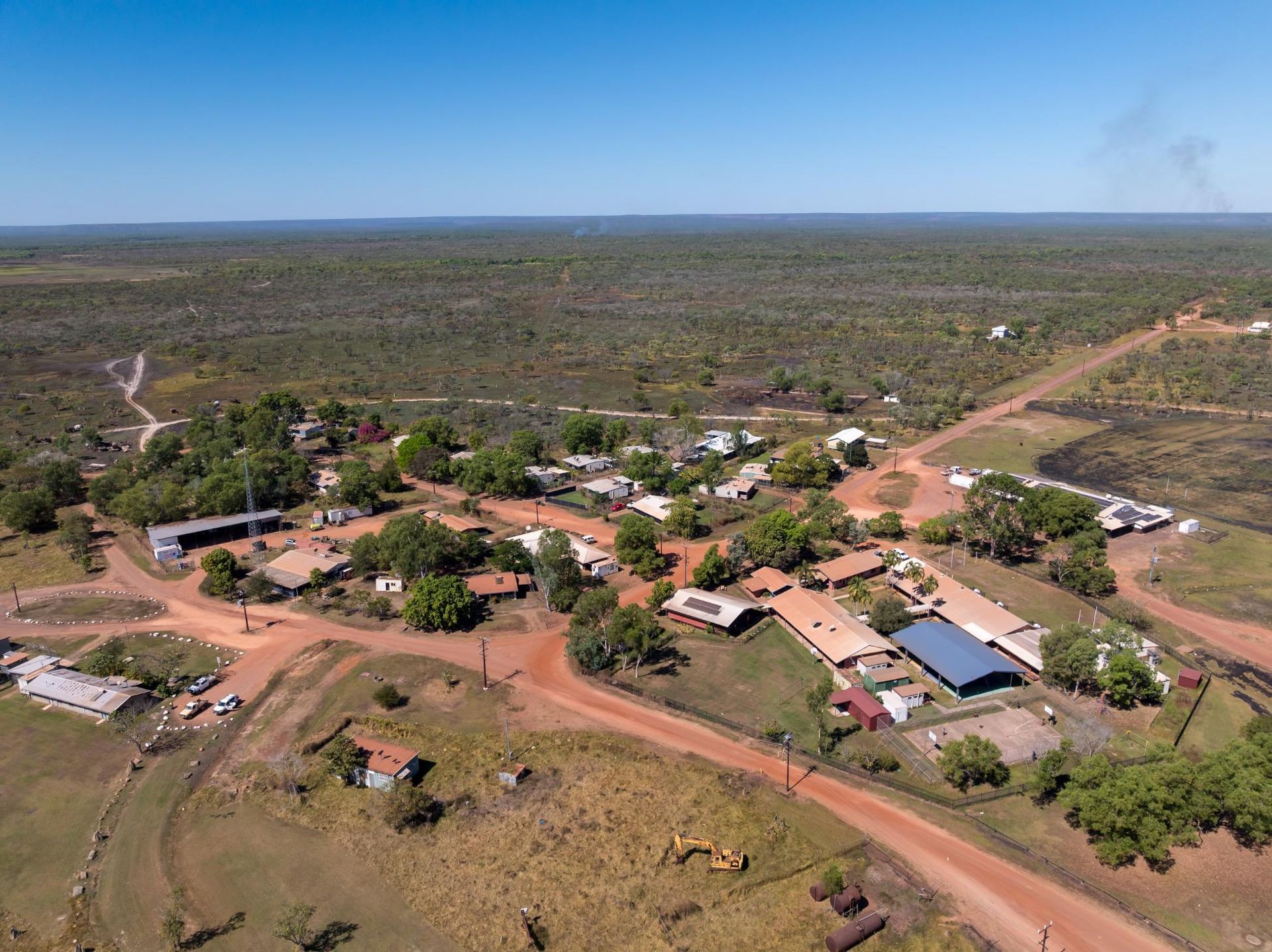 Aerial view of a small rural town with red dirt roads and low buildings surrounded by green trees under a blue sky.