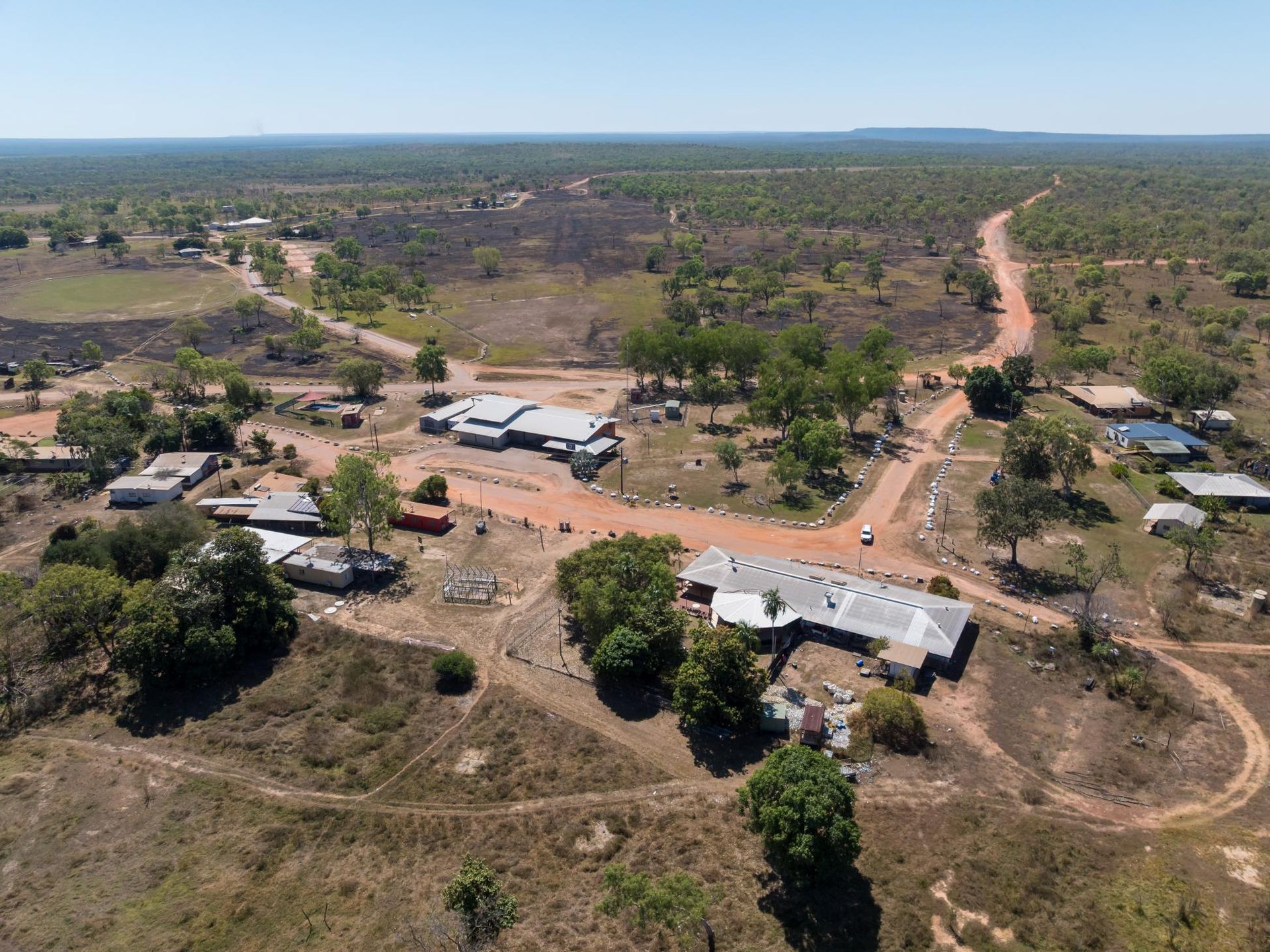 Aerial view of a small rural community with buildings, dirt roads, and surrounding dry landscape under a blue sky.