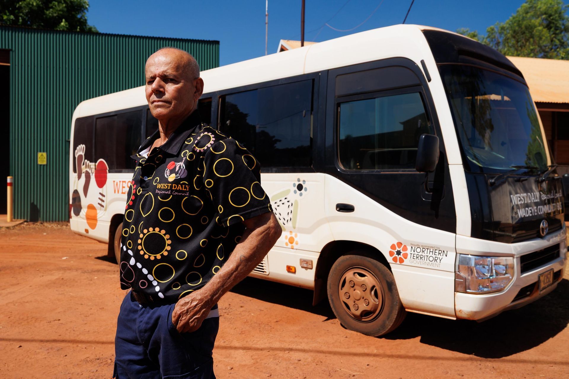 Man stands beside a white bus. The bus has colorful designs on its side, in front of a green building.