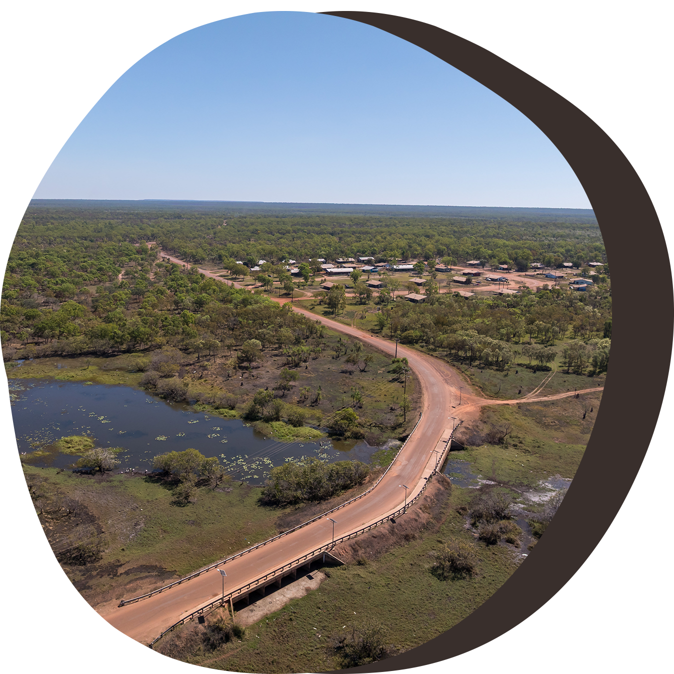 Aerial view of a bridge over wetlands, road leading to a small settlement, surrounded by green trees.