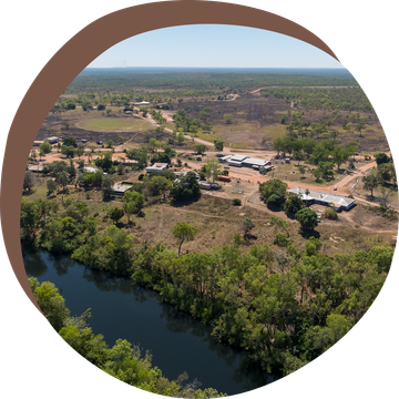 Aerial view of a settlement near a river, surrounded by dry vegetation and a vast landscape under a blue sky.