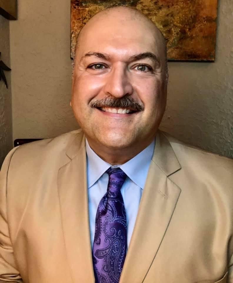 A bald man with a mustache is wearing a tan suit and purple tie.