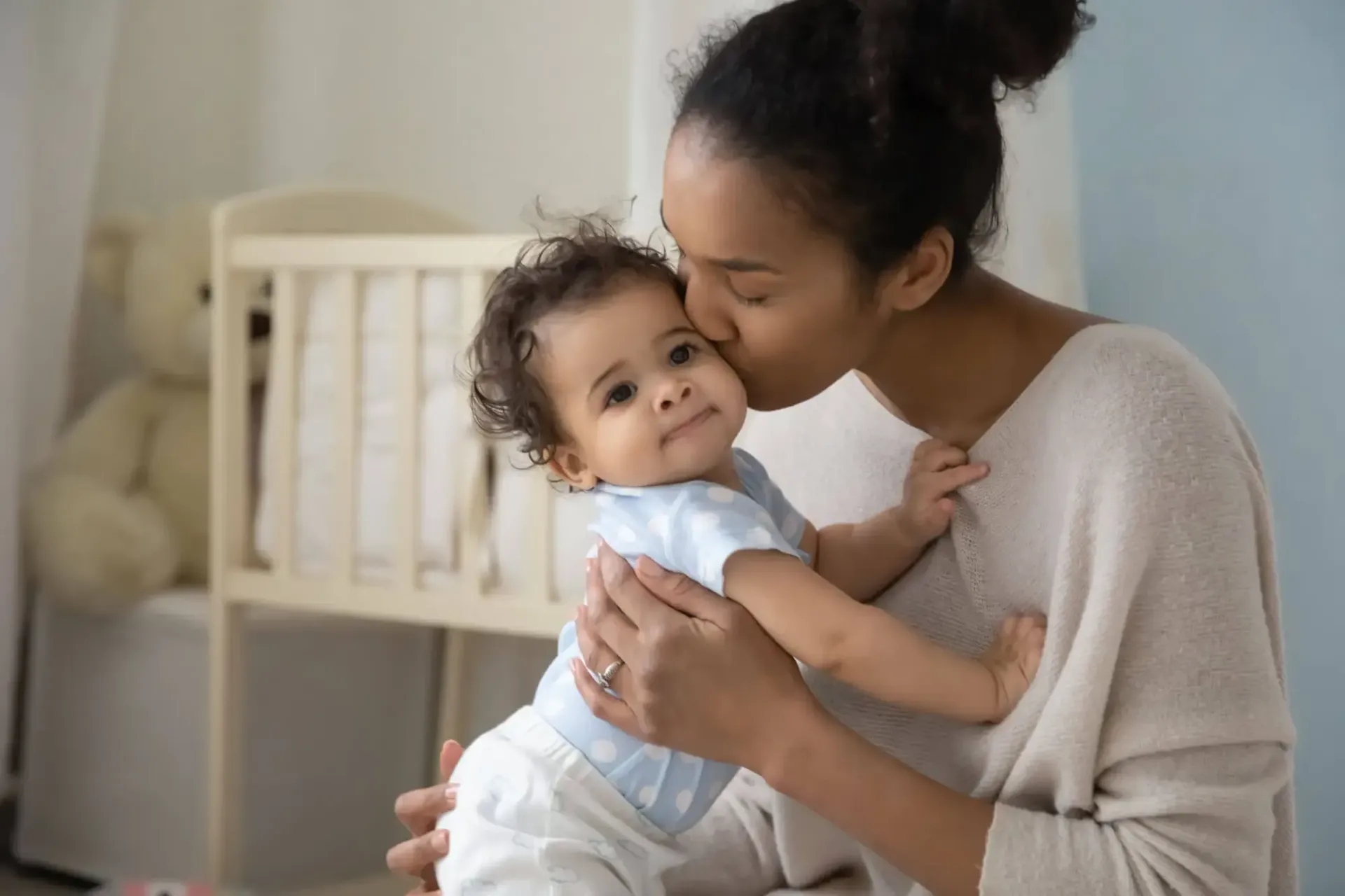 A woman is kissing her baby on the cheek in a nursery.