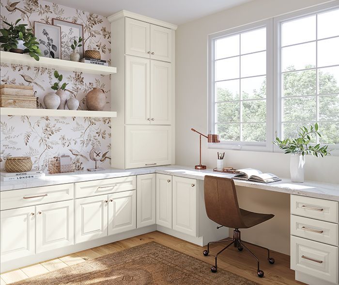 Cream-colored home office with built-in desk, cabinets, and floral wallpaper. A window and brown chair are visible. Cream-colored home office with built-in desk, cabinets, and floral wallpaper. A window and brown chair are visible.