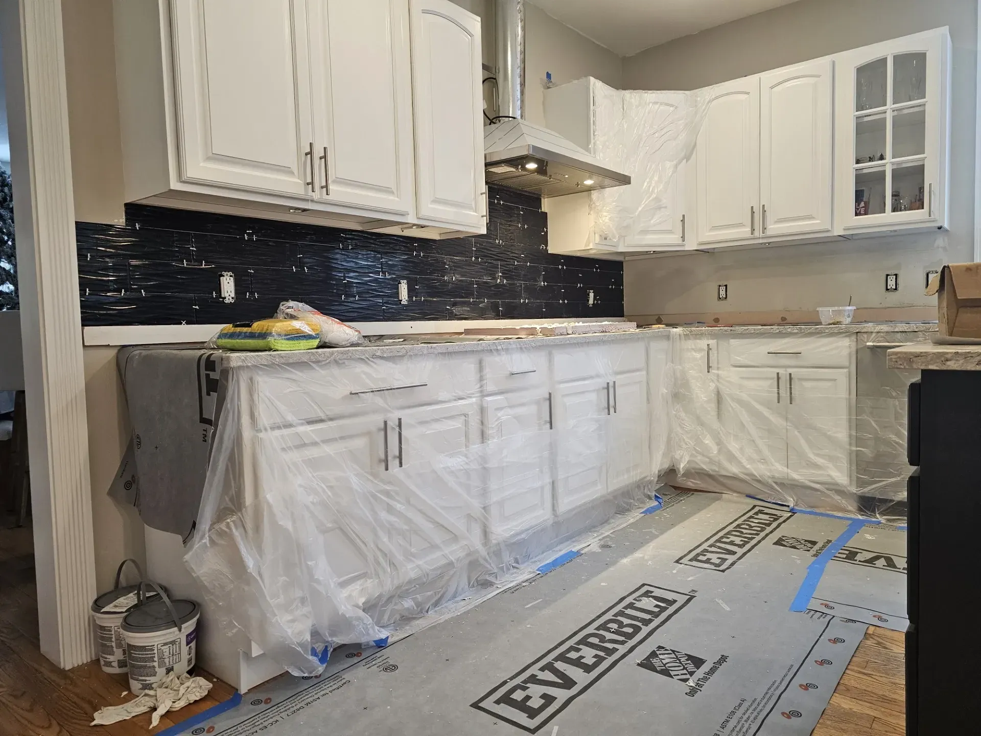 A kitchen under renovation with white cabinets, a dark tile backsplash, and protective plastic covering the counters.