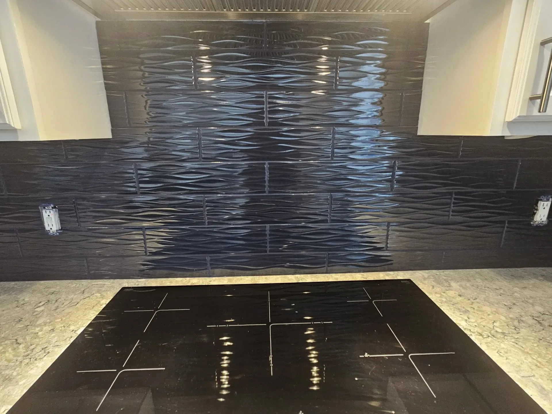 A black textured tile backsplash behind a black electric stovetop and granite countertop.