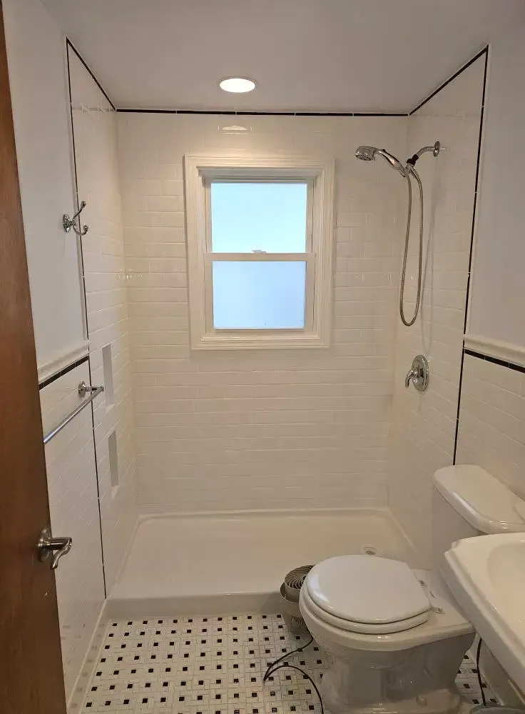 Small, white tiled bathroom with shower, toilet, and window, black and white checkered floor, shower head.