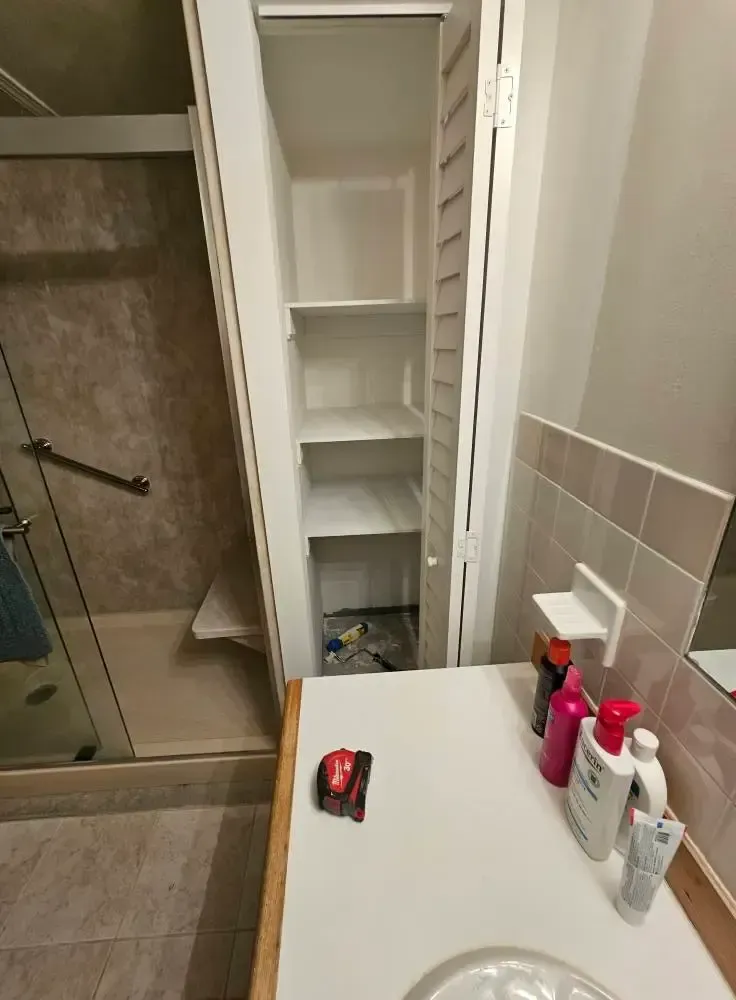 Bathroom with shower, tall cabinet, and sink. White cabinet has open shelves. Sink counter holds toiletries.