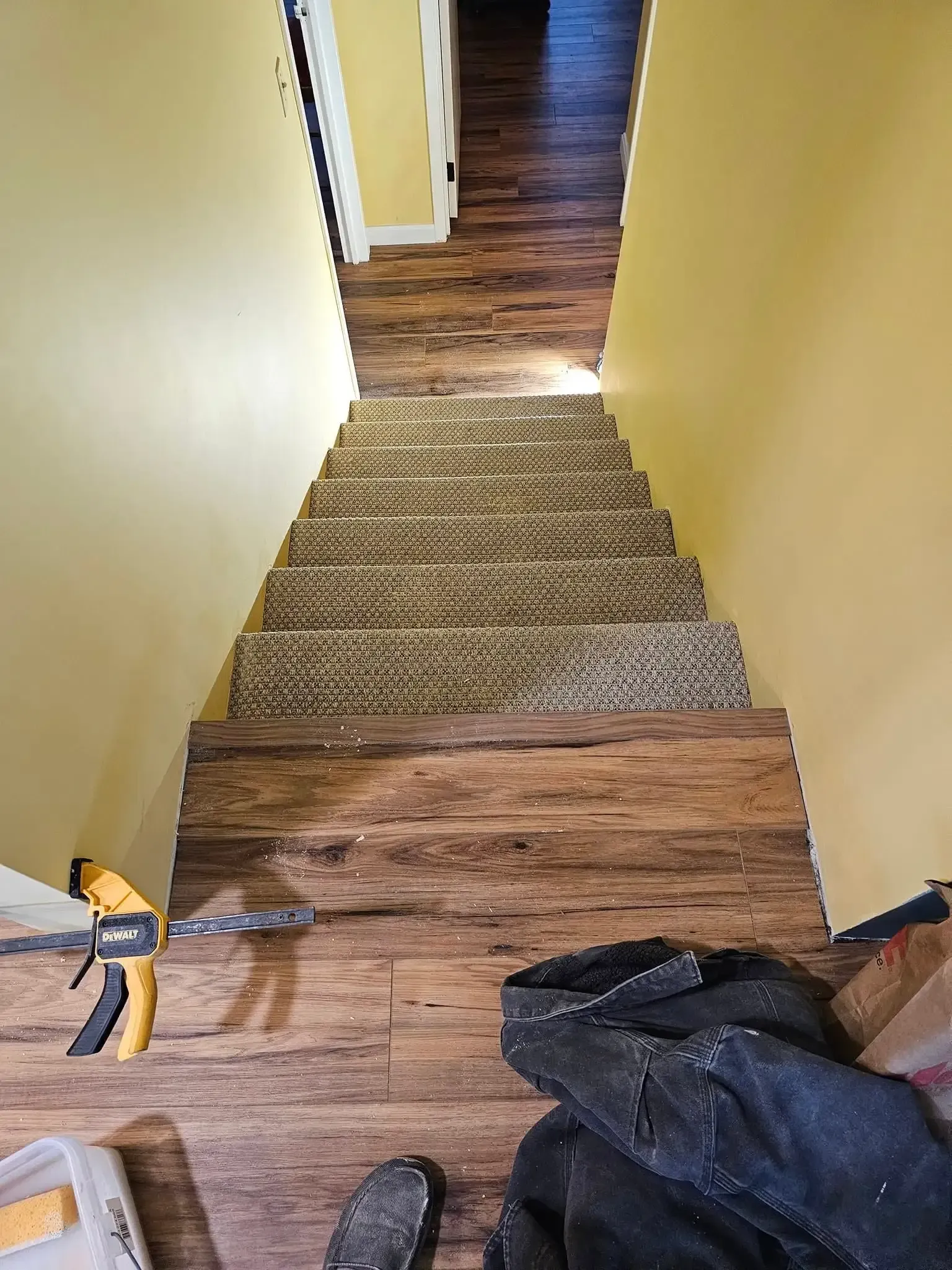 Wooden stairs with brown flooring, cardboard step covers, yellow walls. A tool and clothing are at the bottom.
