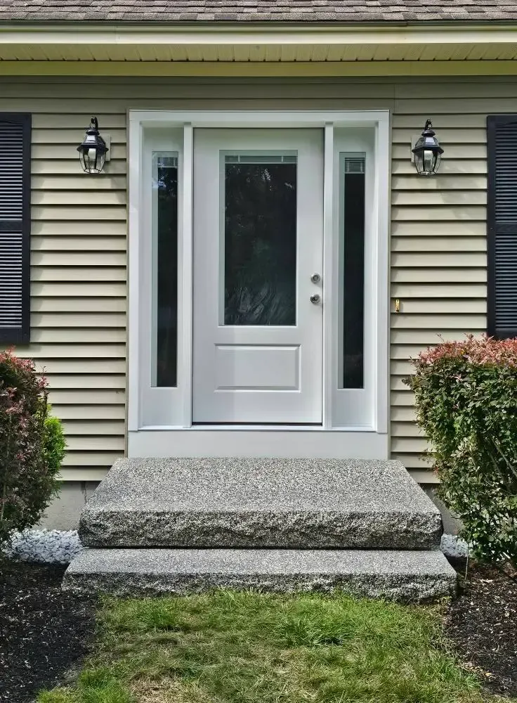 White front door with sidelights and concrete steps. Tan siding, black shutters, and two sconces.